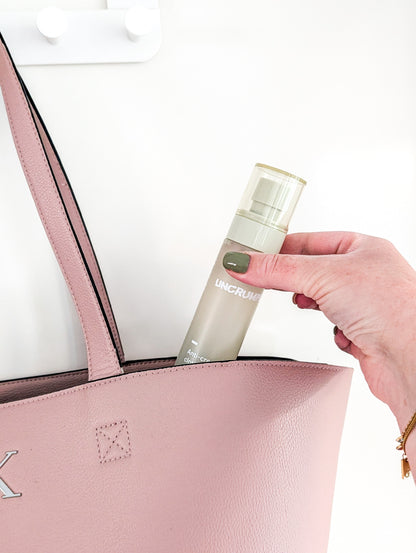 Hand holding a a travel-sized Uncrumpled anti-crease spray bottle, slipping it into a pink handbag on a white background
